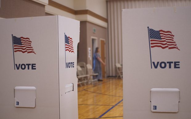 In the space between two voting booths, a woman is seen entering the polling place in the background.