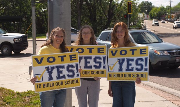 Lisa Easton and her two daughters stand at an intersection holding signs saying "Vote YES to build our school."